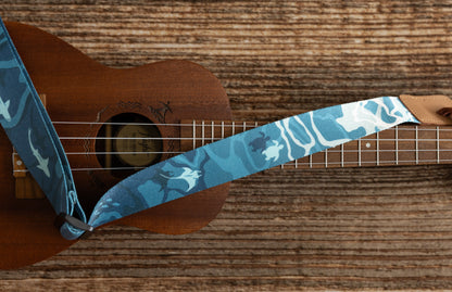 Ocean-designed themed Ukulele Strap. A mix of light and dark blues printed design of ocean water, marine life, turtles, sharks, fish, and manta rays Attached Ukulele lying on wood floor