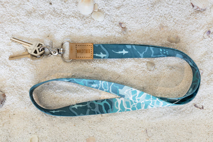 A blue and teal lanyard with a shark and turtle ocean-themed pattern is shown on a sandy surface with scattered seashells. A set of keys hangs from the metal clasp.