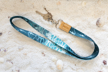 An overhead shot of a blue and teal lanyard with an ocean pattern, including sharks and sea turtles. It's resting on a white sandy surface with seashells, and a pair of keys is attached to the metal clasp.
