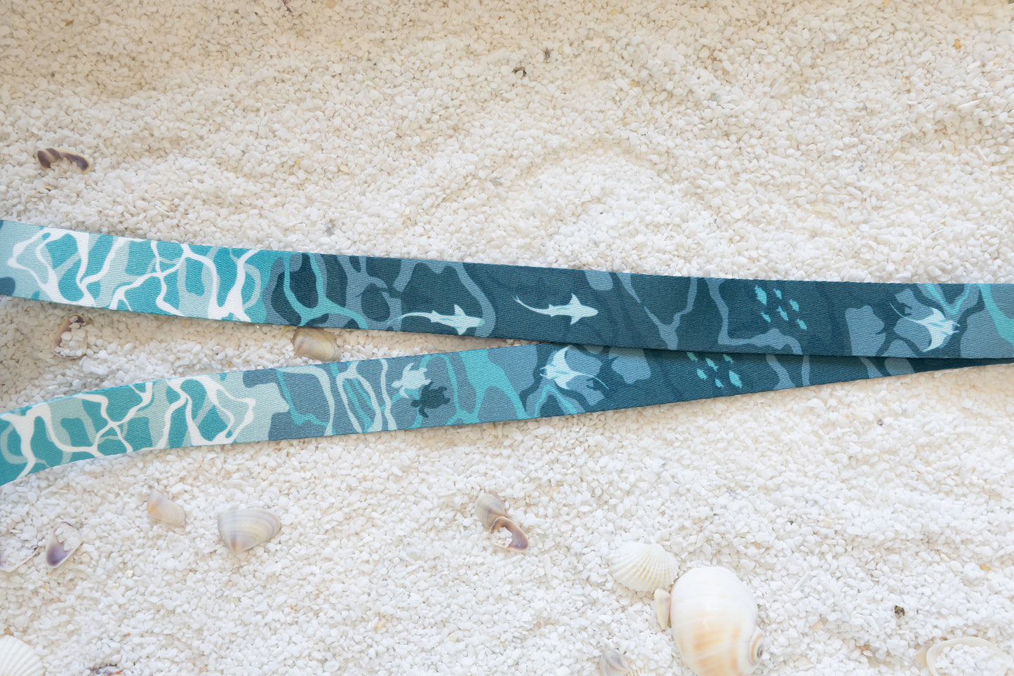A close-up, top-down view of the blue and teal ocean-themed lanyard, laid flat on a sandy surface with seashells. The pattern of sharks and sea turtles is clearly visible.