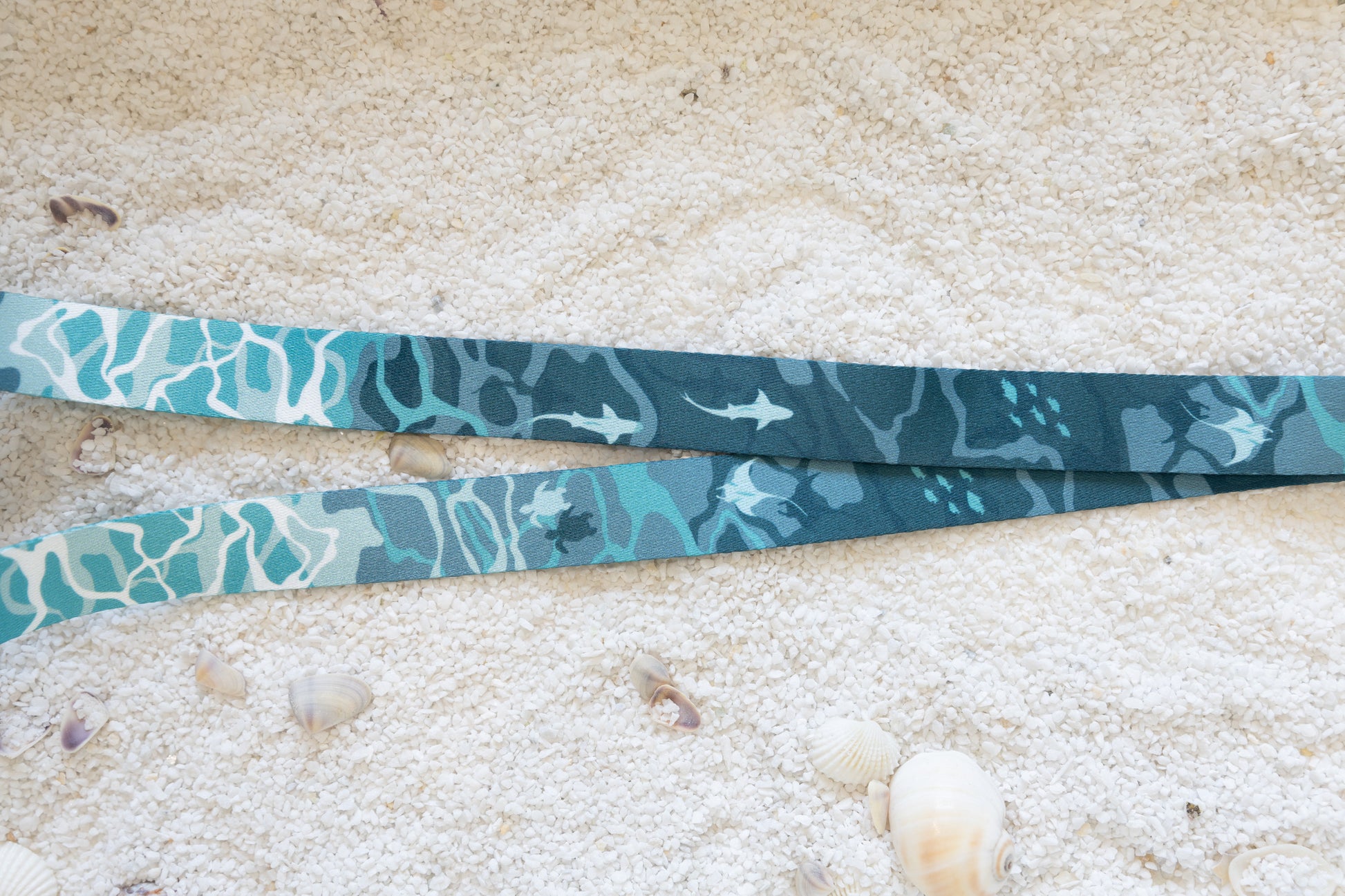 A close-up, top-down view of the blue and teal ocean-themed lanyard, laid flat on a sandy surface with seashells. The pattern of sharks and sea turtles is clearly visible.