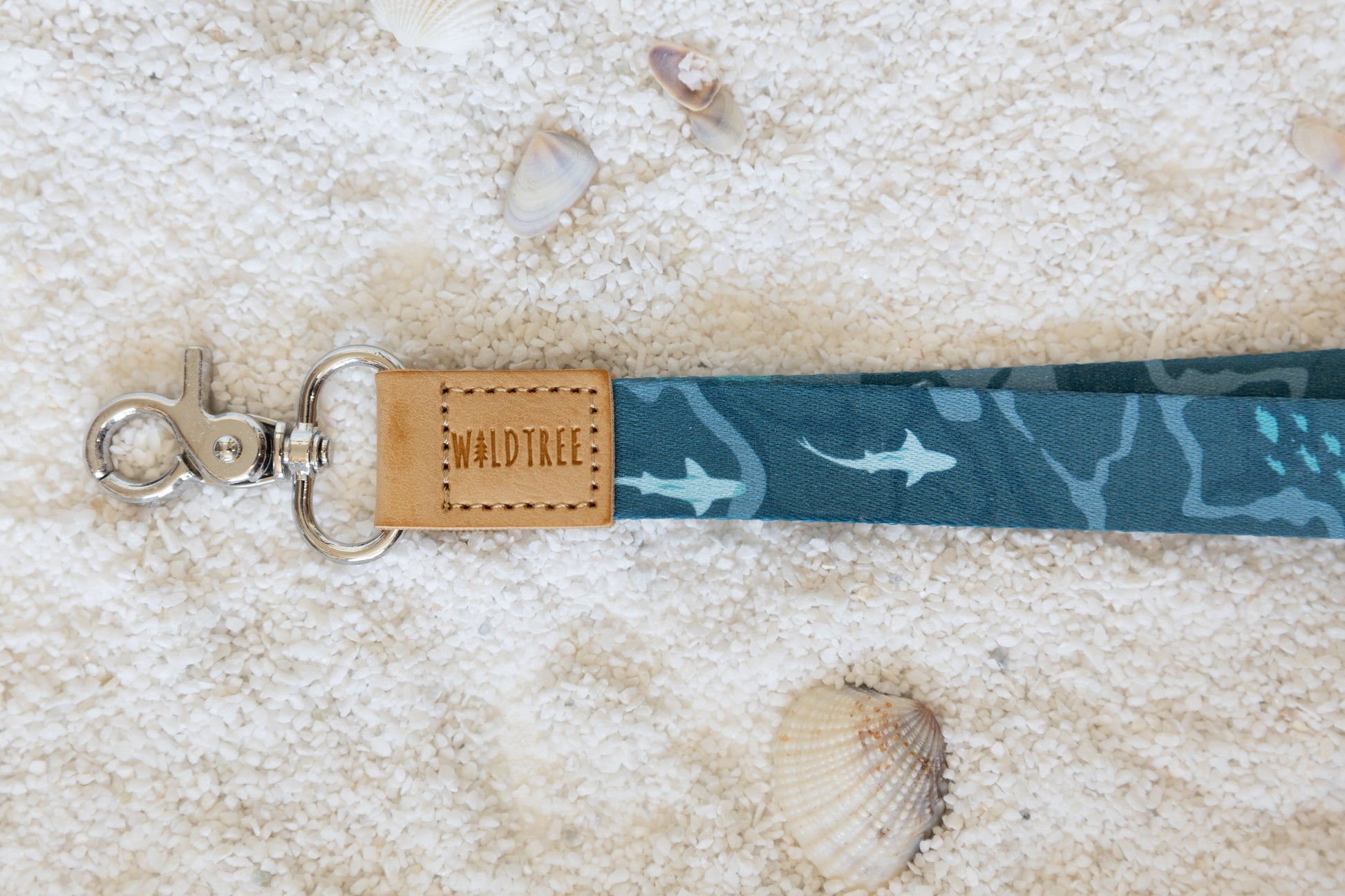 A close-up shot of the blue and teal ocean-themed lanyard, focusing on the tan leather tag that reads "WILDTREE." The metal clasp and part of the pattern, including a shark, are visible. The lanyard is on a sandy surface with a seashell.