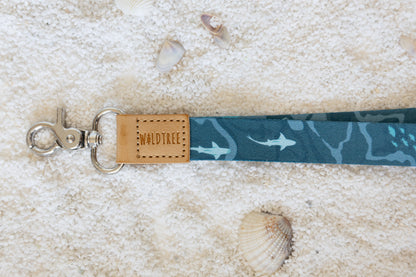 A close-up shot of the blue and teal ocean-themed lanyard, focusing on the tan leather tag that reads "WILDTREE." The metal clasp and part of the pattern, including a shark, are visible. The lanyard is on a sandy surface with a seashell.