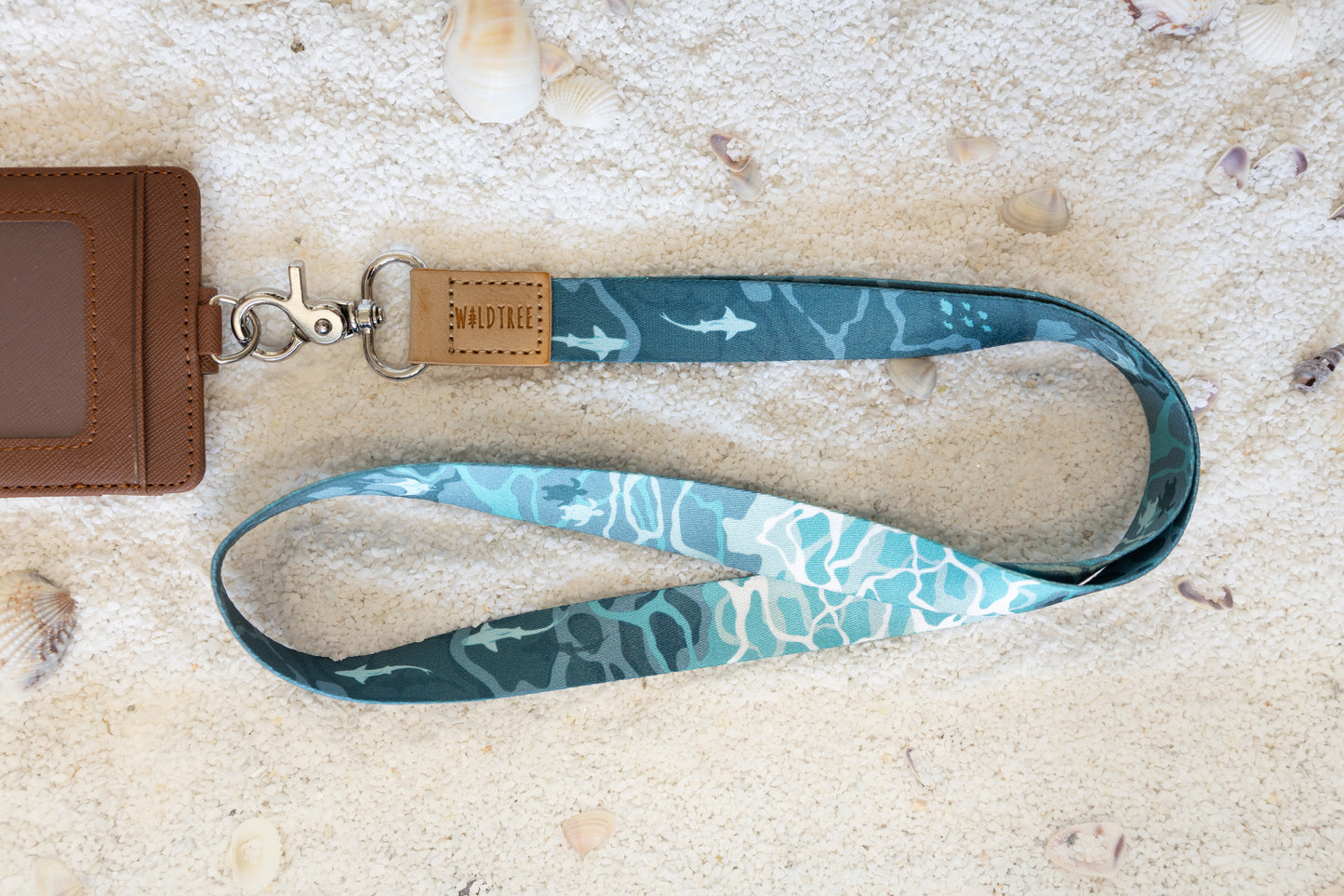 A blue and teal lanyard with a shark and turtle pattern is shown on a sandy surface with seashells. A brown cardholder is partially visible to the left. The lanyard has a tan leather tab with "WILDTREE" embossed on it.