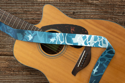 An overhead shot of the guitar body with the strap laid horizontally across the strings, showcasing the gradient from dark teal to white "water ripple" patterns and marine life icons.