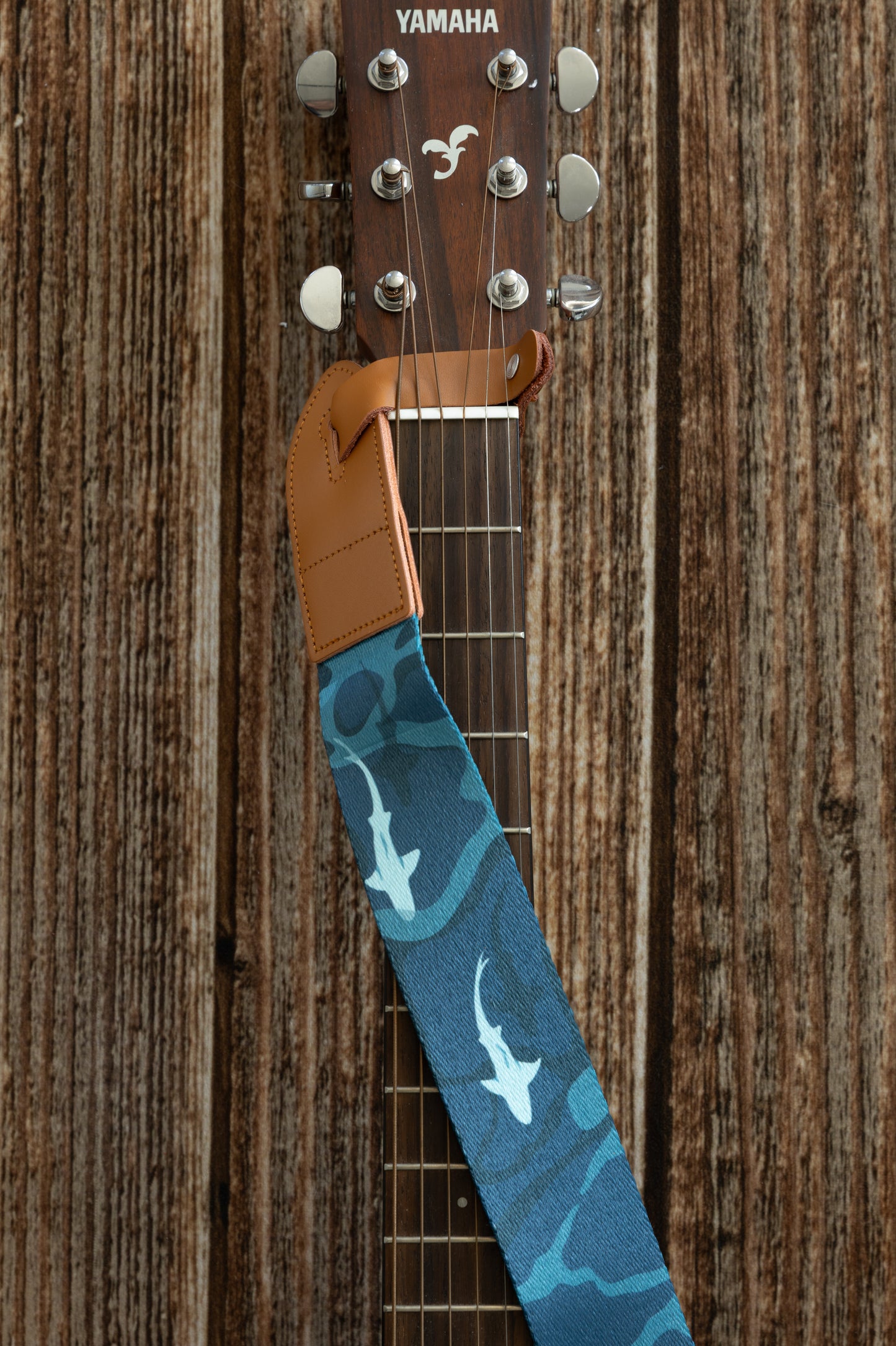 A close-up of the guitar headstock showing the tan leather end of the strap attached to the neck, featuring shark illustrations on the blue fabric below.
