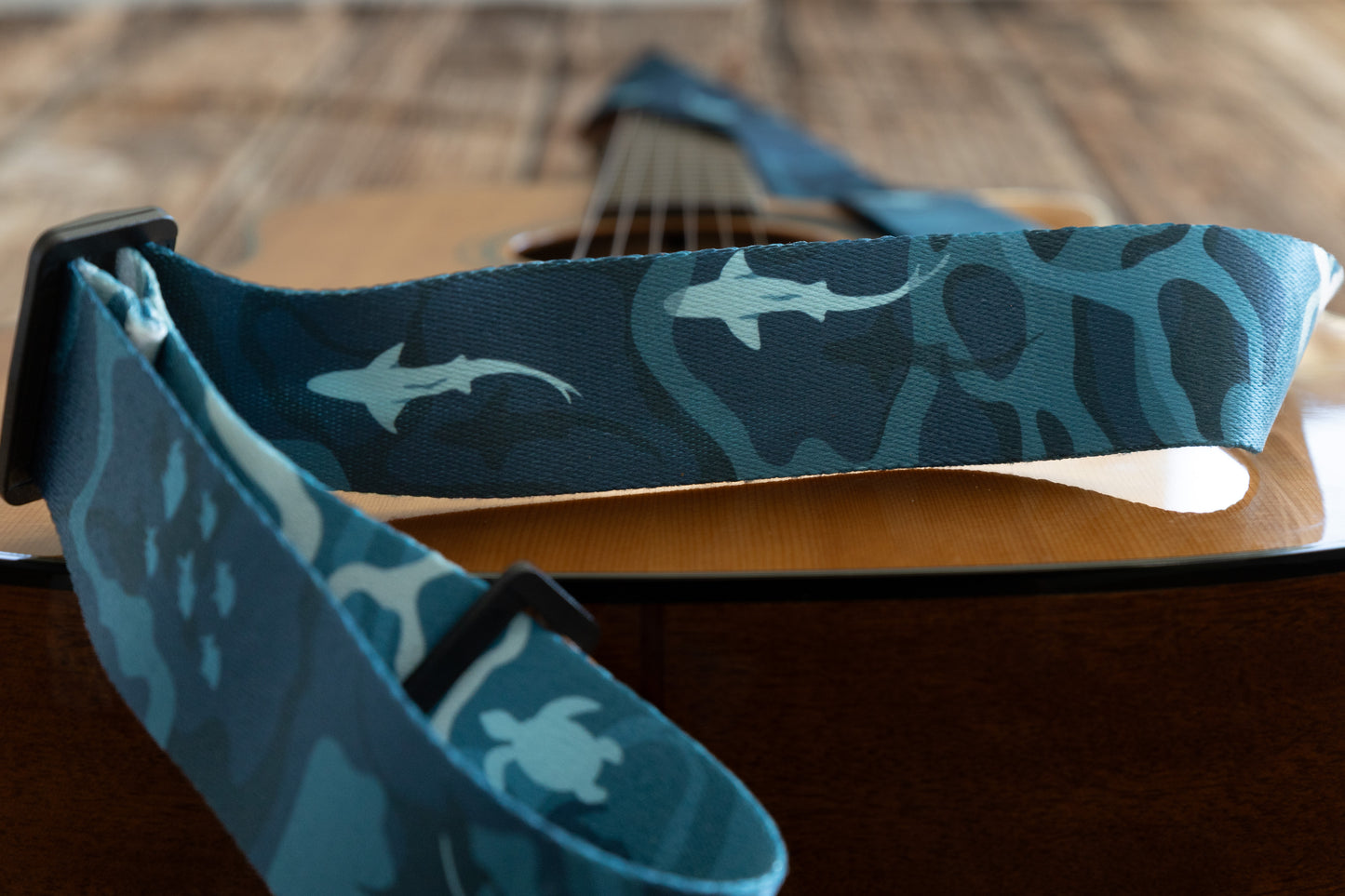 A close-up, low-angle shot of the guitar strap showing light blue shark silhouettes and sea turtles against a dark teal, wavy background.