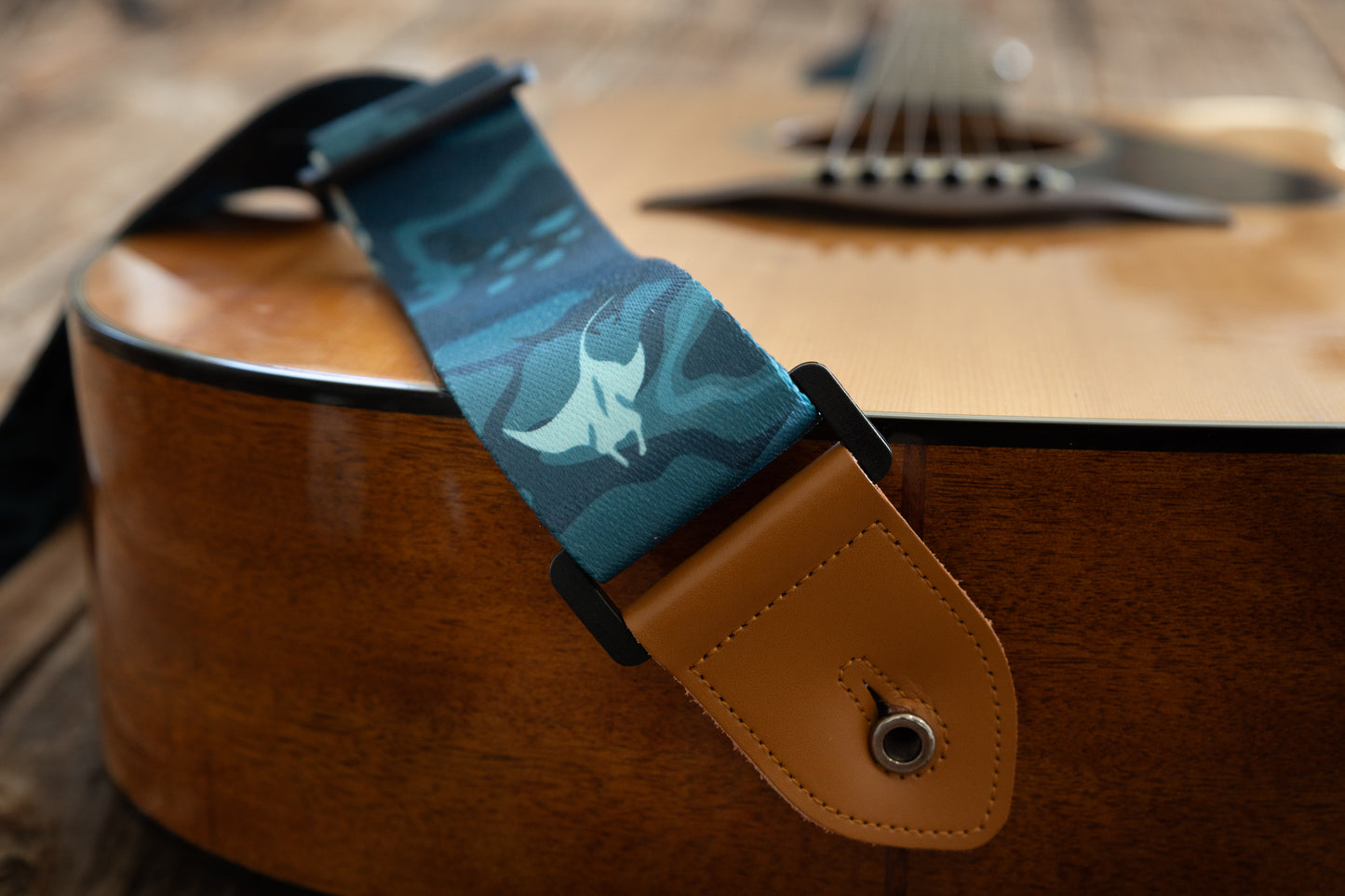 A side-angle detail shot of the guitar strap’s tan leather connector and black buckle resting on the side of the guitar body, with a manta ray design visible on the fabric.