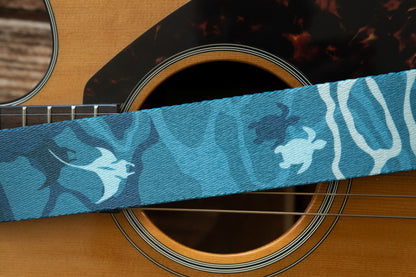 A tight macro shot of the strap passing over the guitar's soundhole, highlighting the texture of the woven fabric and the silhouettes of a manta ray and two sea turtles.
