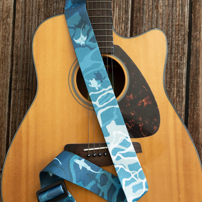 A blue guitar strap featuring a pattern of manta rays, sea turtles, and sharks in various shades of teal and white, draped over the body of an acoustic guitar.