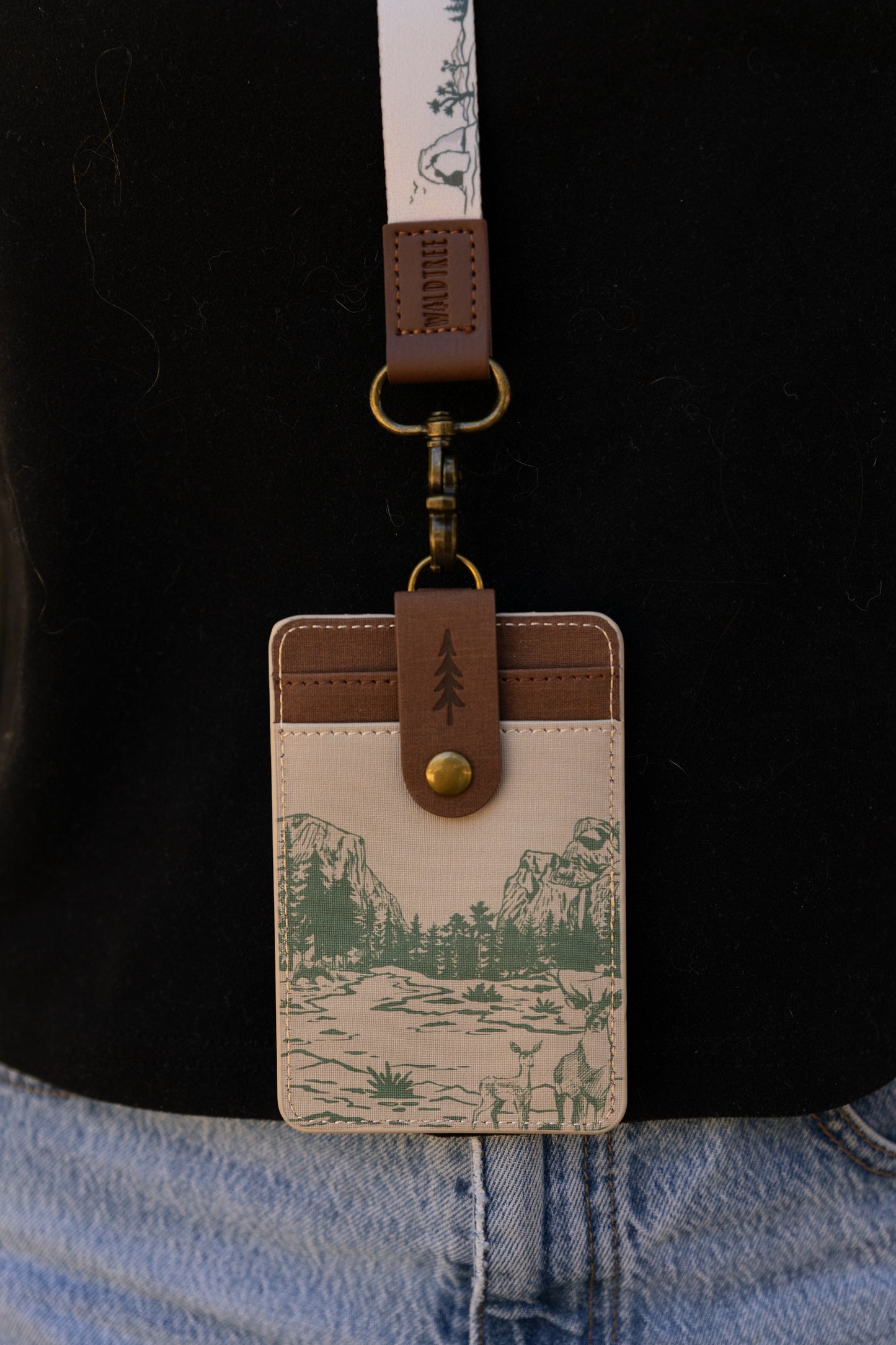 A waist-up view of a person outdoors wearing the cream Yosemite landscape ID holder clipped to a matching patterned lanyard. The image highlights the complete matching set paired casually with a black tee and denim shorts.