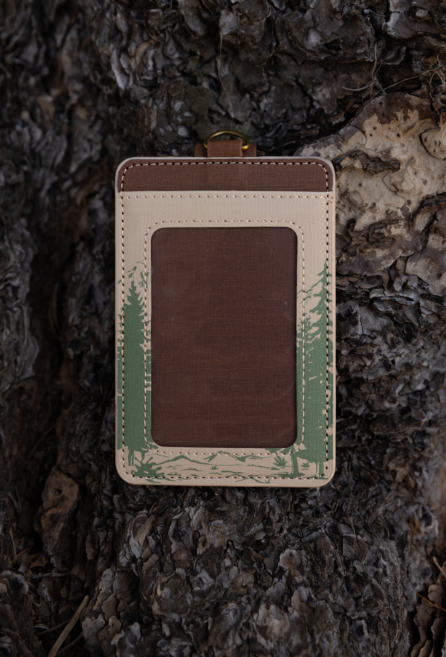 A detailed close-up showing the front clear plastic window frame of the cream-colored Yosemite ID wallet. The detailed stitching, top brass hardware attachment, and green landscape sketches are clearly visible against a wood dark background.
