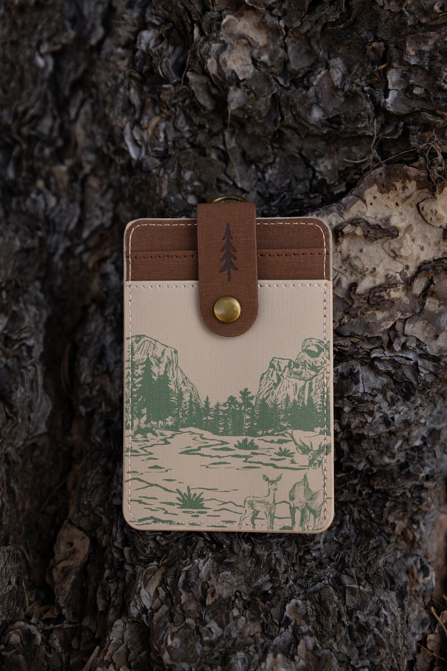 A detailed close-up showing theback side f of the cream-colored Yosemite ID wallet. The detailed stitching, top brass hardware attachment, and green landscape sketches are clearly visible against a dark wood background.