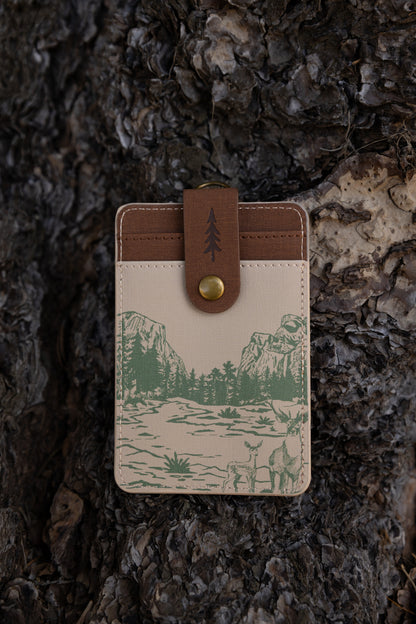 A detailed close-up showing theback side f of the cream-colored Yosemite ID wallet. The detailed stitching, top brass hardware attachment, and green landscape sketches are clearly visible against a dark wood background.