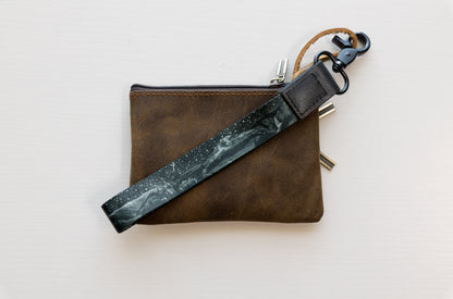 The Parks at Night wristlet keychain attached to a set of keys and draped across a brown leather zip pouch on a white wood surface, showing the black strap printed with mountain silhouettes and a starry night sky.