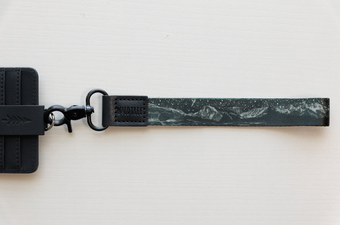 The Parks at Night wristlet keychain attached to a black vegan-leather card holder laid flat on a white background, showing the black strap printed with mountain silhouettes, scattered stars, and a shooting star, and the black swivel clasp.