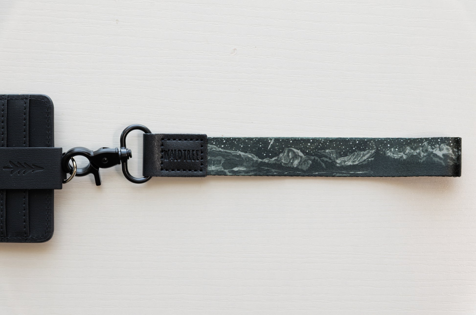 The Parks at Night wristlet keychain attached to a black vegan-leather card holder laid flat on a white background, showing the black strap printed with mountain silhouettes, scattered stars, and a shooting star, and the black swivel clasp.