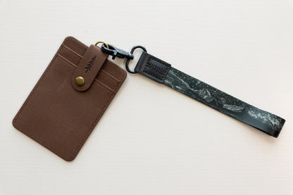 The Parks at Night wristlet keychain attached to a brown vegan-leather card holder on a white background, showing the black strap printed with mountain silhouettes and a starry night sky, and the black leather WILDTREE tab.