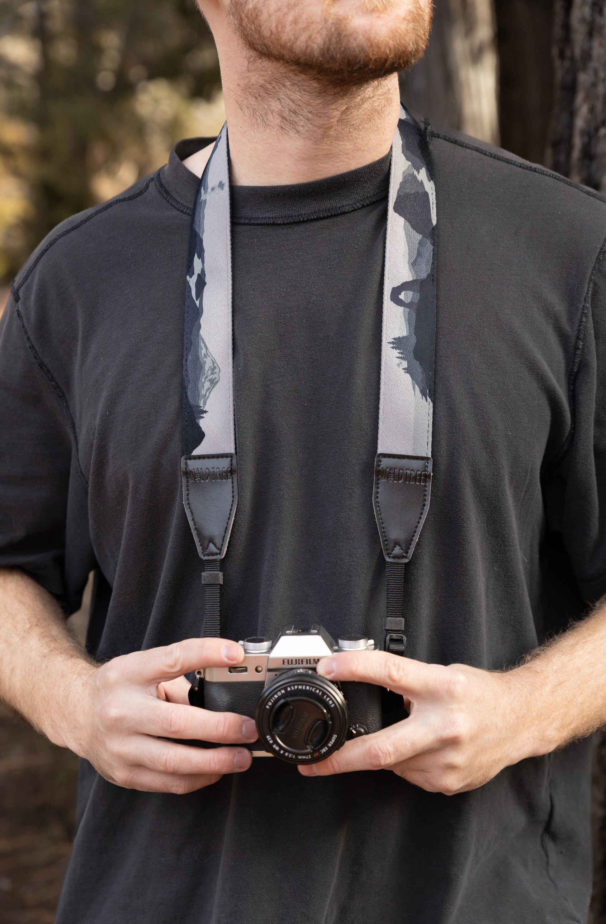 A man wearing a dark grey t-shirt holds a silver Fujifilm camera. The camera is secured by a wide strap featuring a black, white, and grey mountain landscape print, finished with black leather tabs embossed with the WILDTREE logo.
