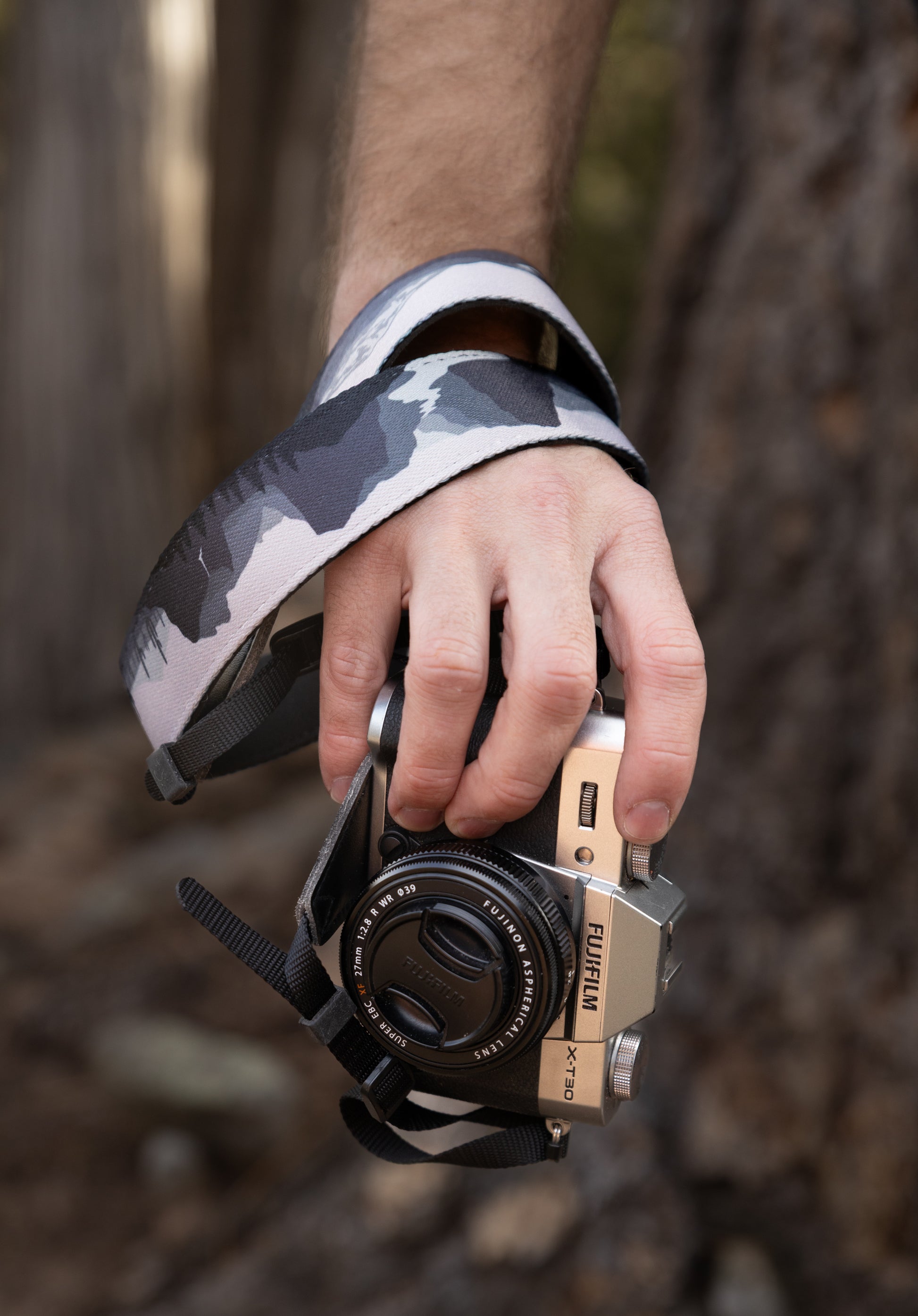 A close-up shot of a hand holding a silver Fujifilm camera with the WILDTREE "Parks in Black and White" strap wrapped securely around the wrist. The strap displays a high-contrast mountain and forest graphic in grayscale.