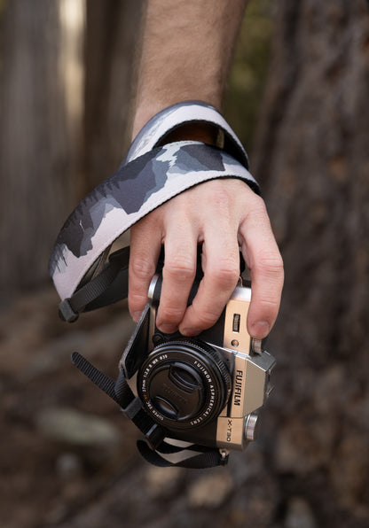 A close-up shot of a hand holding a silver Fujifilm camera with the WILDTREE "Parks in Black and White" strap wrapped securely around the wrist. The strap displays a high-contrast mountain and forest graphic in grayscale.