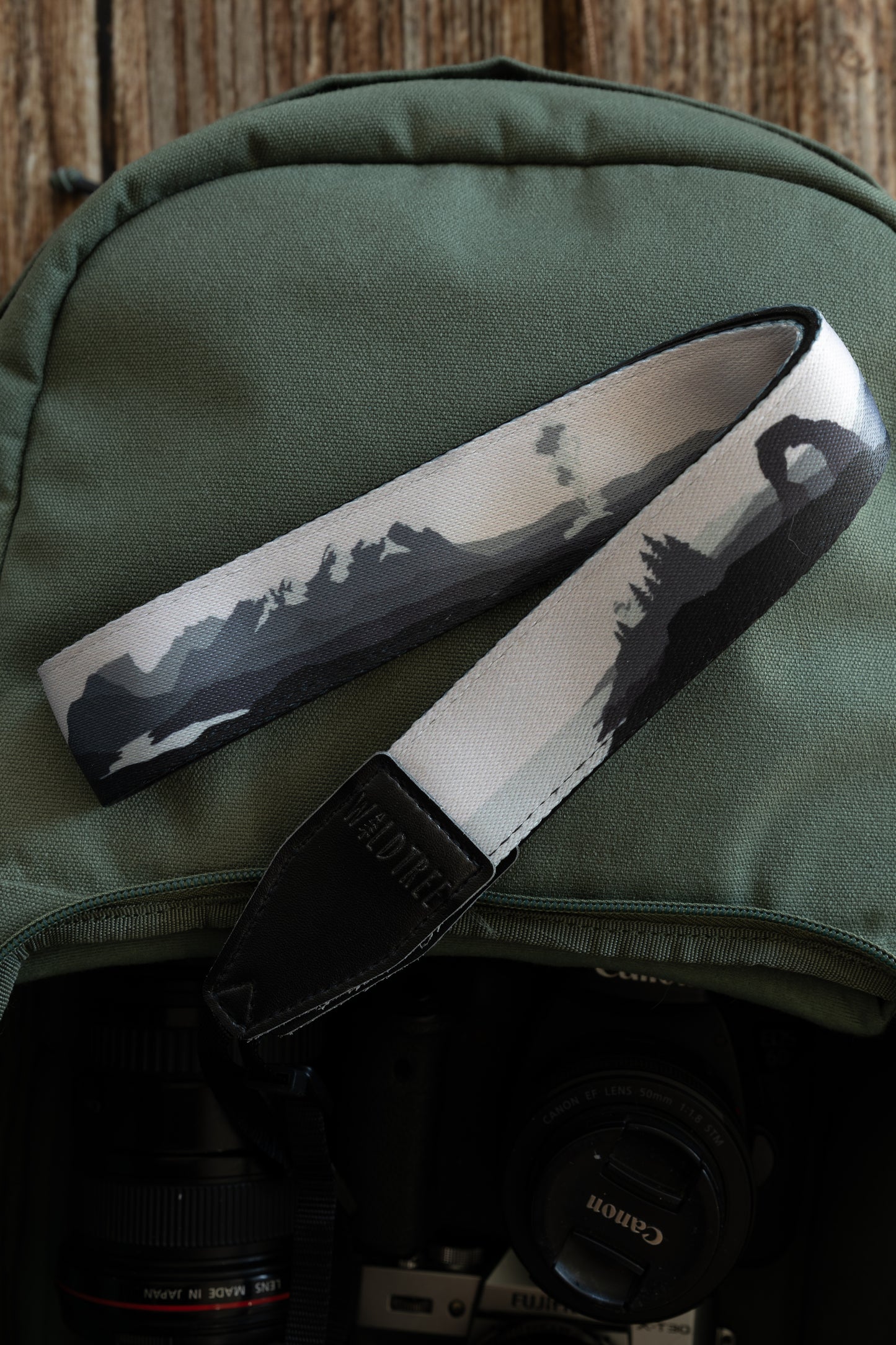 A camera strap with a monochromatic grayscale illustration of national park landscapes, including mountain peaks and the Delicate Arch, draped over a green canvas backpack. A camera lens is visible in the bag below.