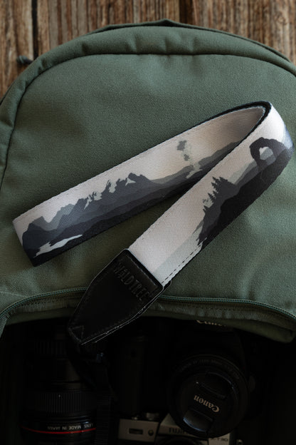 A camera strap with a monochromatic grayscale illustration of national park landscapes, including mountain peaks and the Delicate Arch, draped over a green canvas backpack. A camera lens is visible in the bag below.