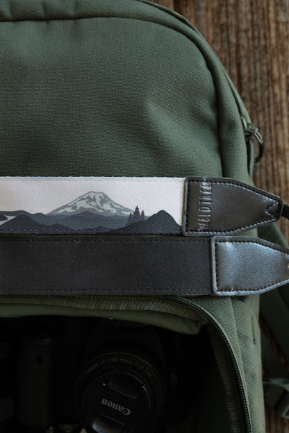 A close-up of two camera straps resting on a green backpack. The top strap features a grayscale illustration of a snow-capped mountain (Mount Rainier) and evergreen trees, while the bottom strap shows its solid black, textured underside. Both have black leather ends embossed with "WILDTREE."
