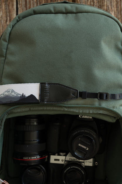 A wide shot showing a camera strap with a mountain landscape design resting across the middle of a green backpack. The backpack is open at the bottom, revealing a Canon DSLR, a Fujifilm mirrorless camera, and multiple lenses neatly packed inside.