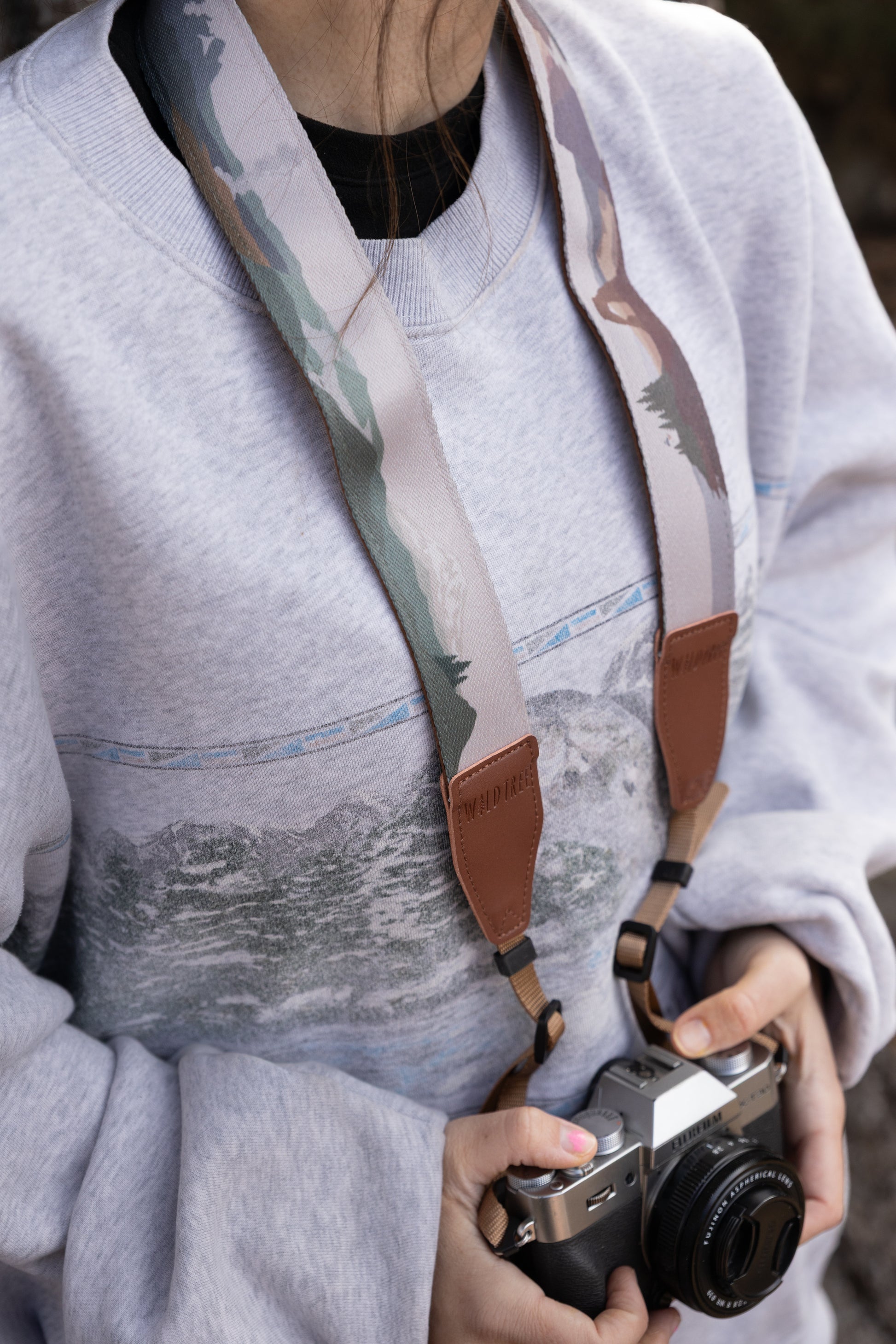 A close-up of a person wearing a grey sweatshirt with a mountain graphic, holding a silver Fujifilm camera. The WILDTREE "Parks in Color" strap is draped around their neck, showing a soft brown leather underside and tan connection cords.