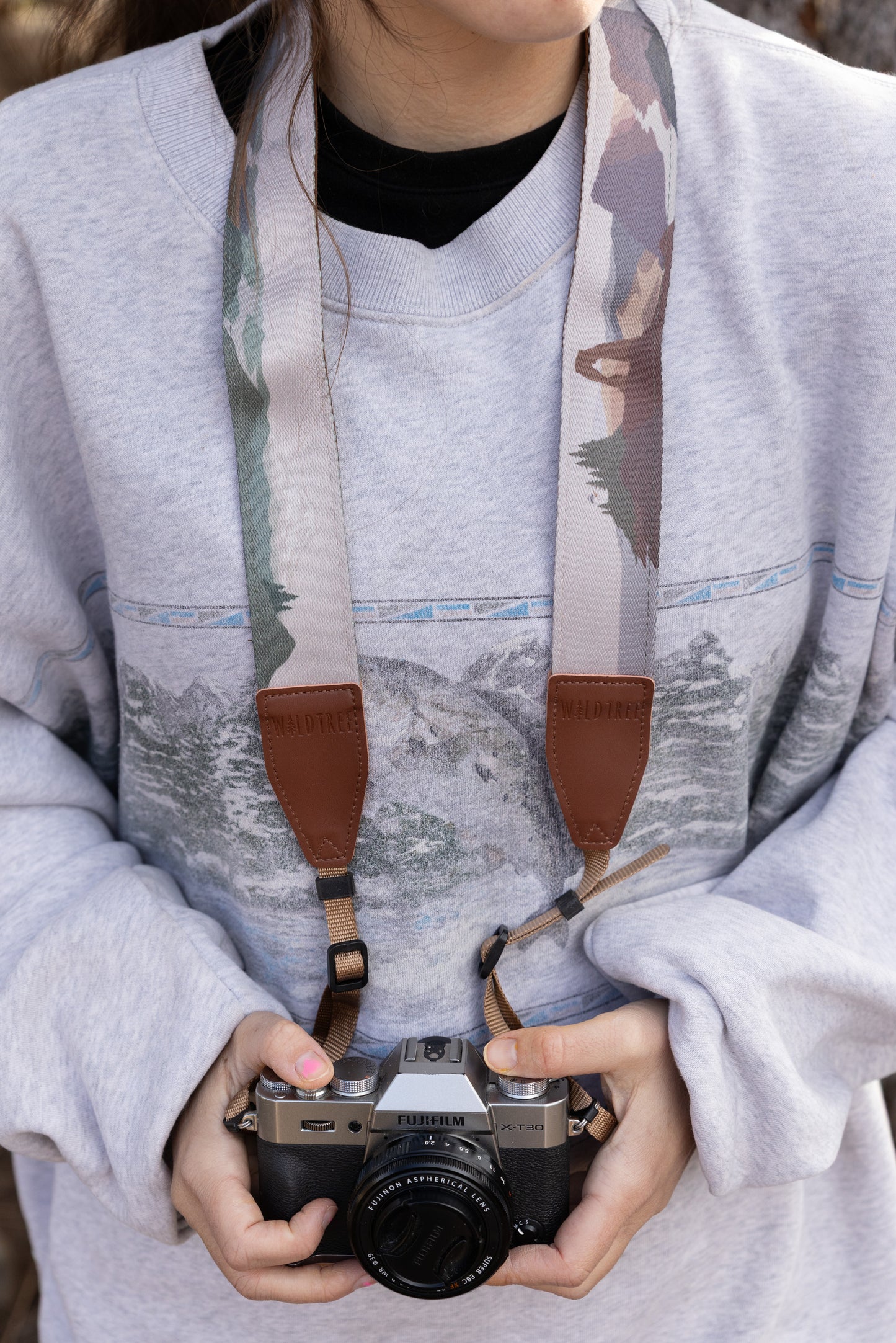 A medium shot of a person in a grey sweatshirt holding a Fujifilm camera at waist level. The colorful National Park strap is clearly displayed, showing two brown leather WILDTREE tabs and the variety of landscape scenes printed on the webbing.