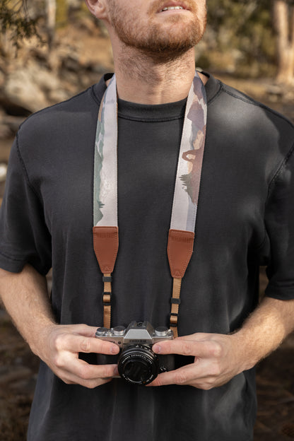 A front view of a man in a black t-shirt wearing a camera strap around his neck. The strap features colorful, stylized illustrations of National Park landscapes, including a desert arch. It is secured with brown leather end-tabs embossed with the WILDTREE logo.