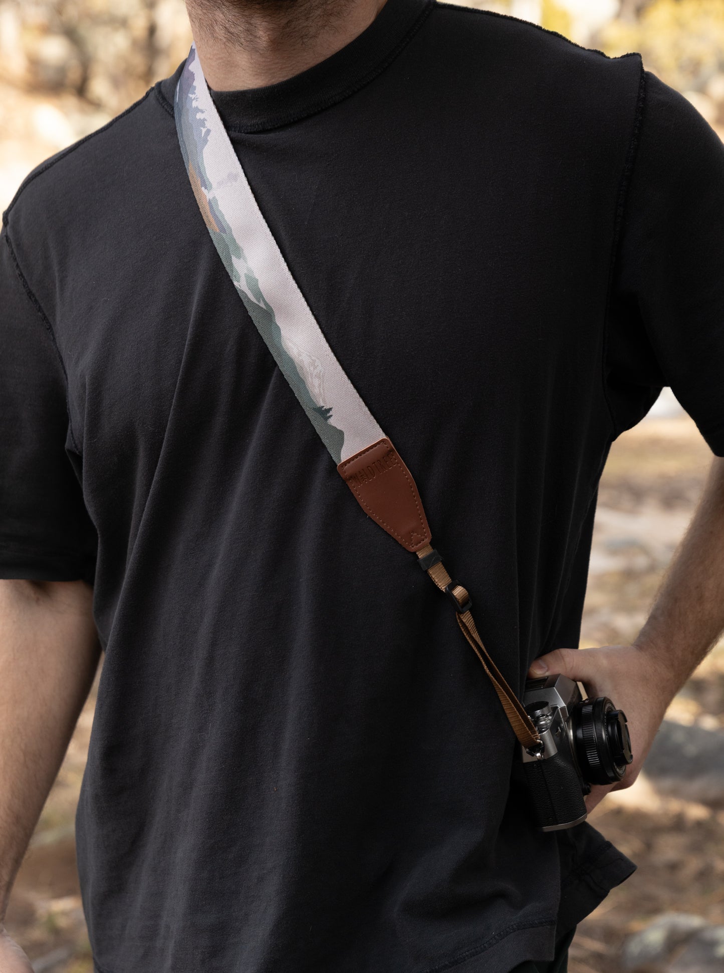 A side view of a man wearing a black t-shirt with a colorful National Park-themed camera strap worn across his body as a sling. The brown WILDTREE leather tab is visible at the chest level as the camera rests at his hip.