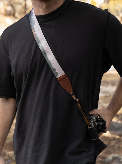 A side view of a man wearing a black t-shirt with a colorful National Park-themed camera strap worn across his body as a sling. The brown WILDTREE leather tab is visible at the chest level as the camera rests at his hip.