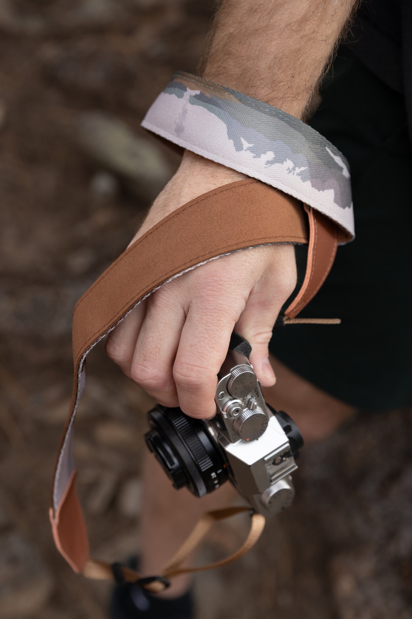 A close-up shot showing a hand holding a silver camera with the WILDTREE "Parks in Color" strap wrapped around the wrist. This view highlights the contrast between the illustrated landscape on the front and the smooth, solid brown backing of the strap.