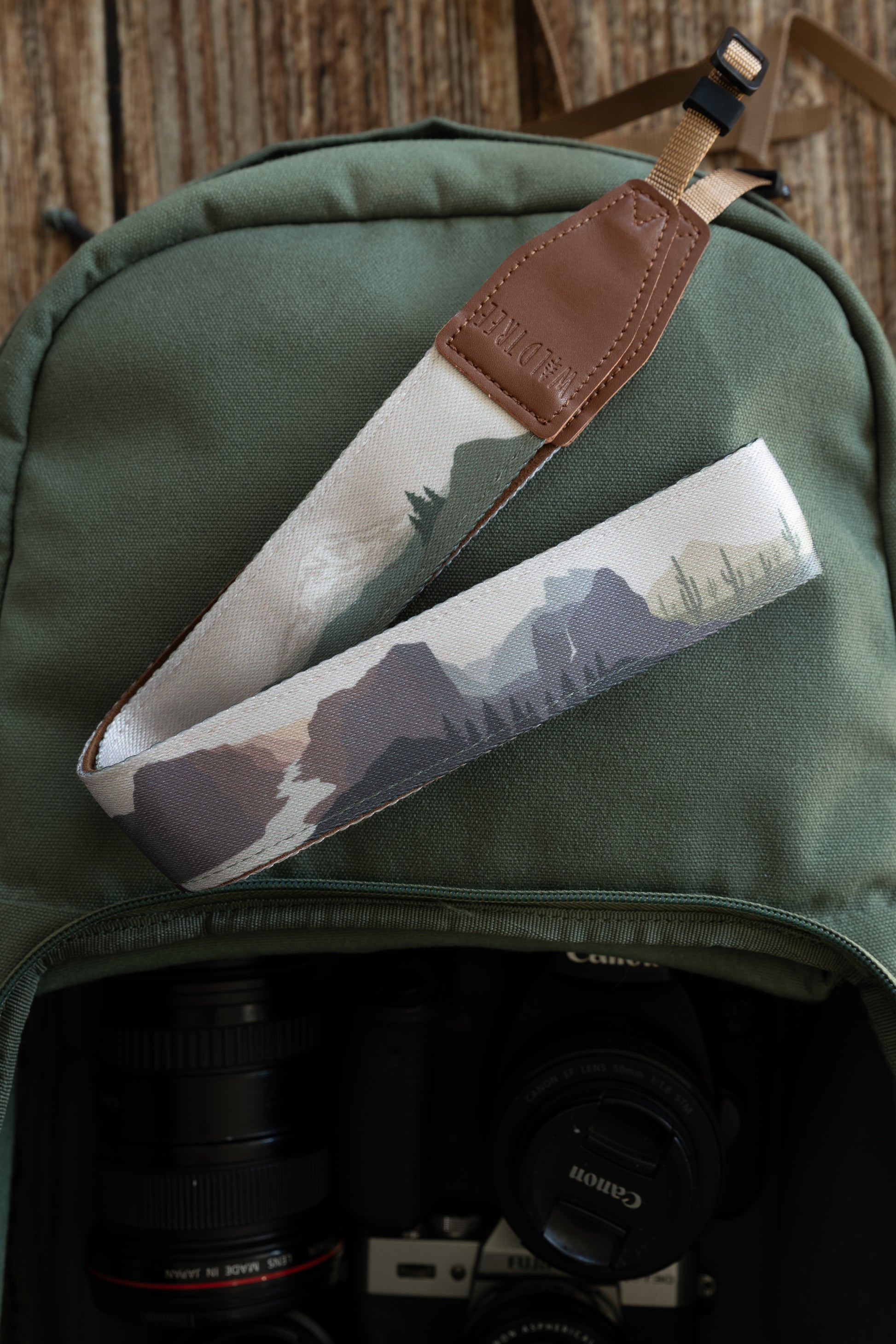 A camera strap with a colorful illustration of national park landscapes—including desert cacti and purple-toned mountains—is draped over a green canvas backpack. The strap features a brown leather end embossed with "WILDTREE" and a tan attachment cord.