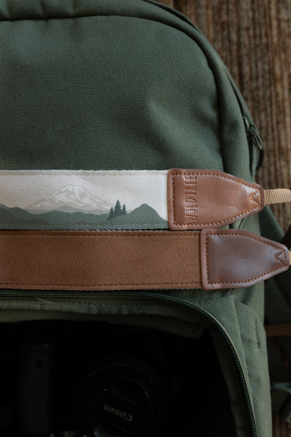 A close-up detail shot of two camera straps resting on a green backpack. The top strap shows a landscape of muted green mountains and a snow-capped peak, finished with a brown leather end. The strap below it shows a solid, soft brown underside.