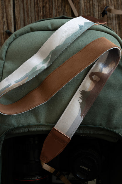 An overhead view of a colorful national parks camera strap coiled on a green backpack. The design transitions between green forest scenes and desert rock arches. A camera and lenses are visible inside the open bag below the strap.