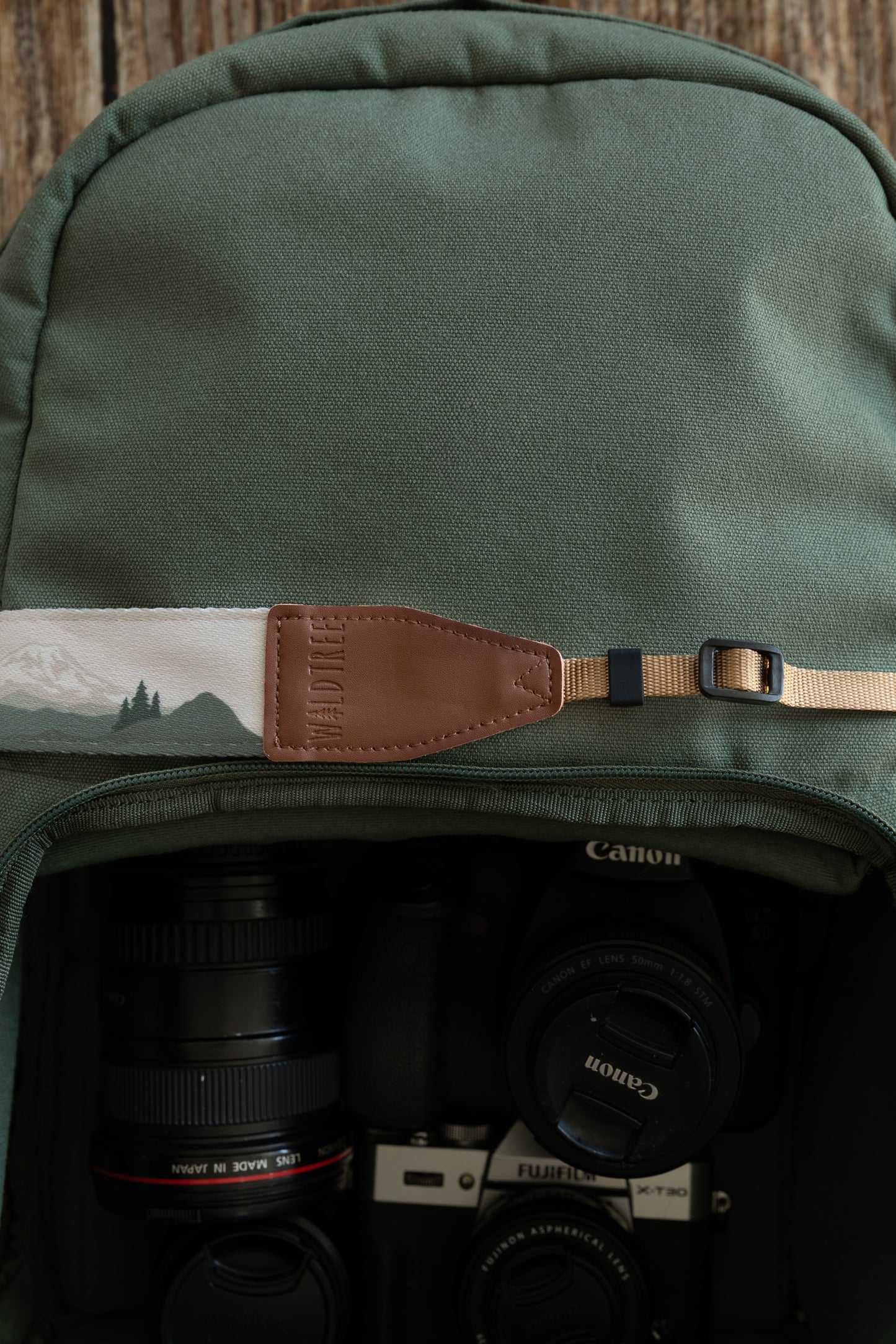 A landscape-style camera strap with a green mountain and forest design laid horizontally across a green backpack. The brown leather end connects to a tan adjustable nylon cord. Several cameras and lenses are organized in the bag's compartment below.