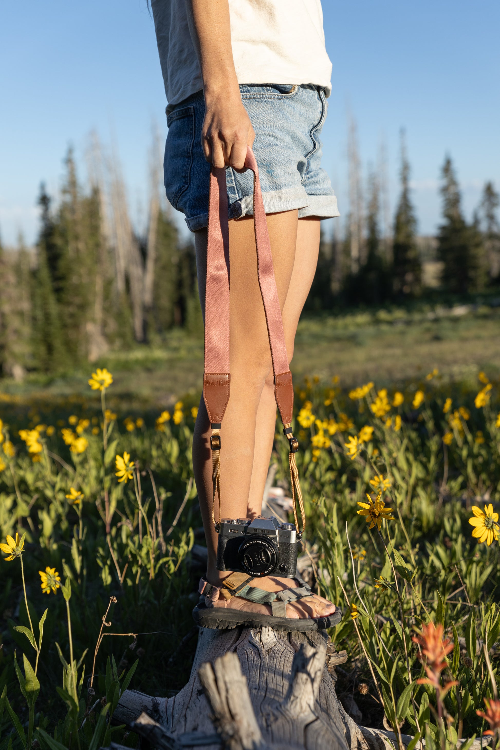 A person in a white shirt and denim shorts stands on a weathered log in a mountain meadow full of bright yellow flowers. They hold a Wildtree Outdoor camera strap featuring a solid muted-pink/rose-colored fabric section connected to a brown leather end piece, with a black and silver camera hanging below.