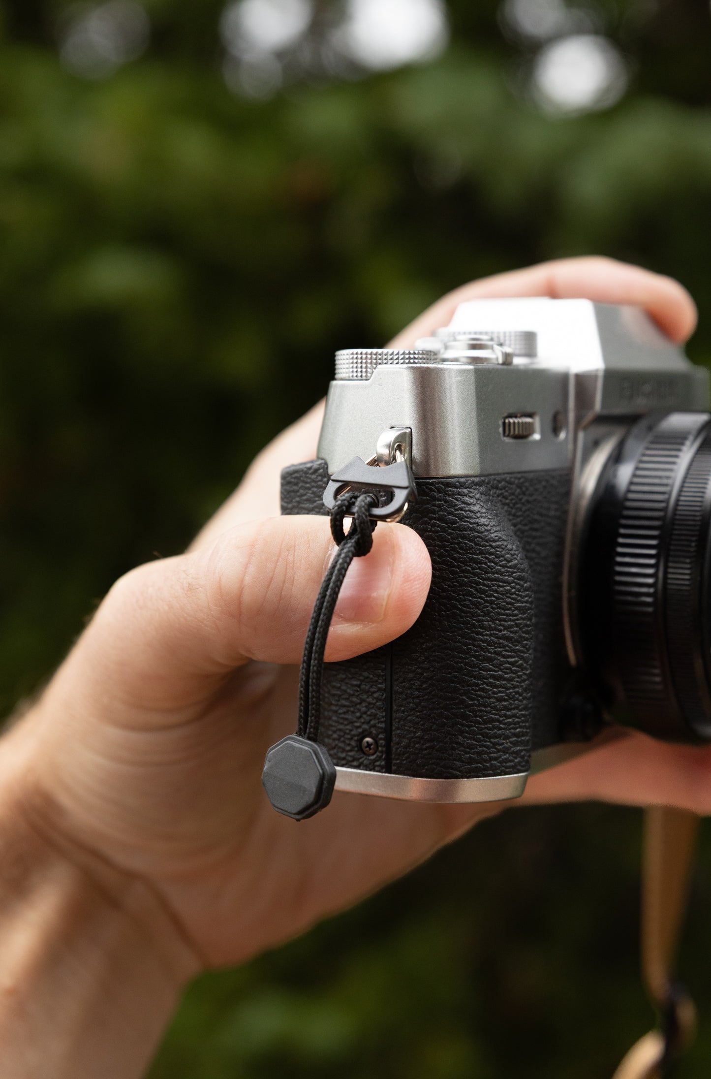 Close-up of a hand holding a silver mirrorless camera with a split-ring anchor and quick-link paracord pull attached to the strap lug
