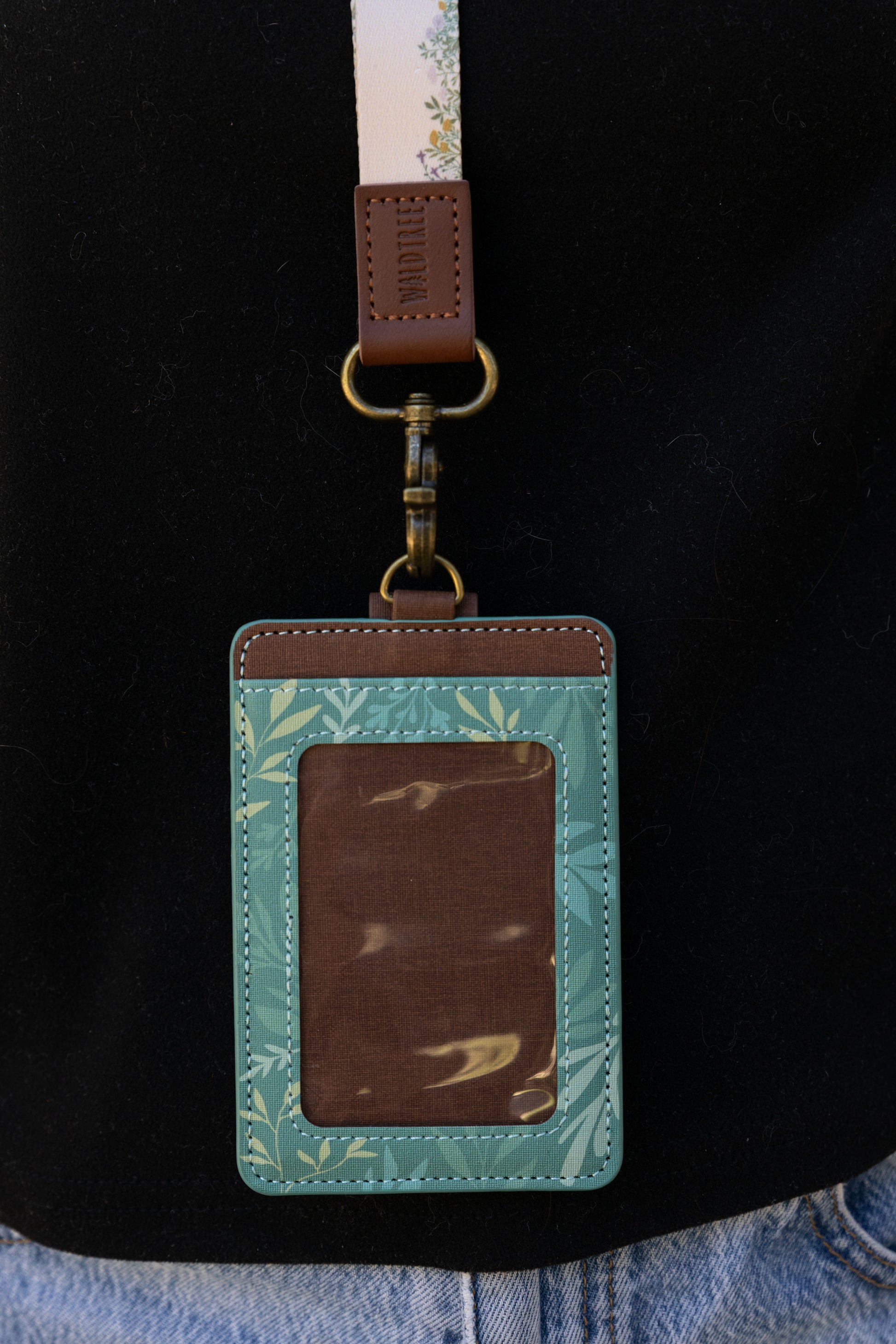 A tight close-up showing the front of the green floral ID holder and its clear plastic badge window. The detailed white stitching, brown trim, and antique brass attachment point are prominently featured.