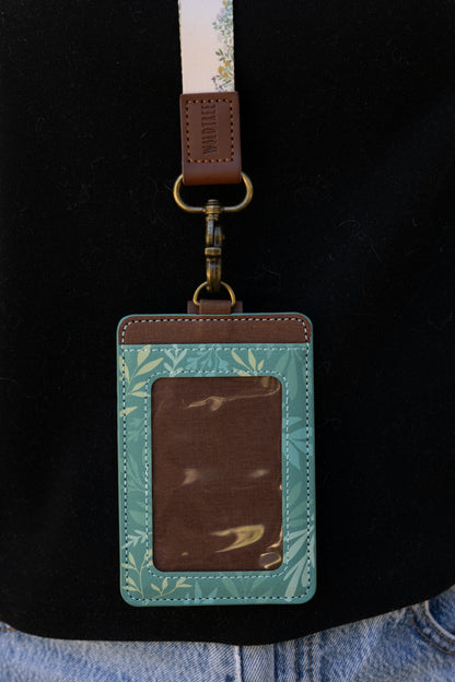 A tight close-up showing the front of the green floral ID holder and its clear plastic badge window. The detailed white stitching, brown trim, and antique brass attachment point are prominently featured.