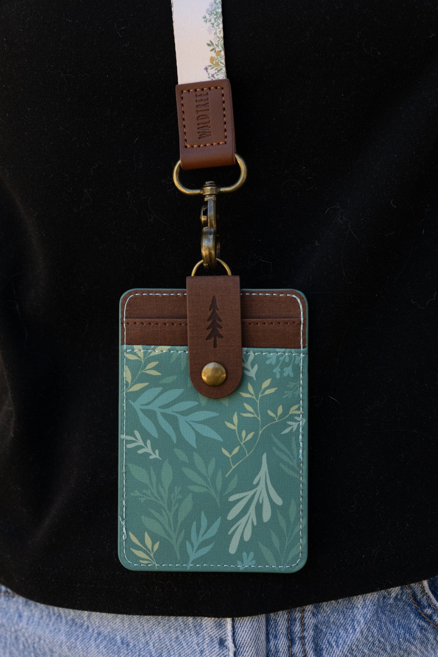 A close-up shot of the back of the green floral ID holder attached to a lanyard. It highlights the brown faux-leather snap strap with a debossed pine tree icon that secures the extra card slots.
