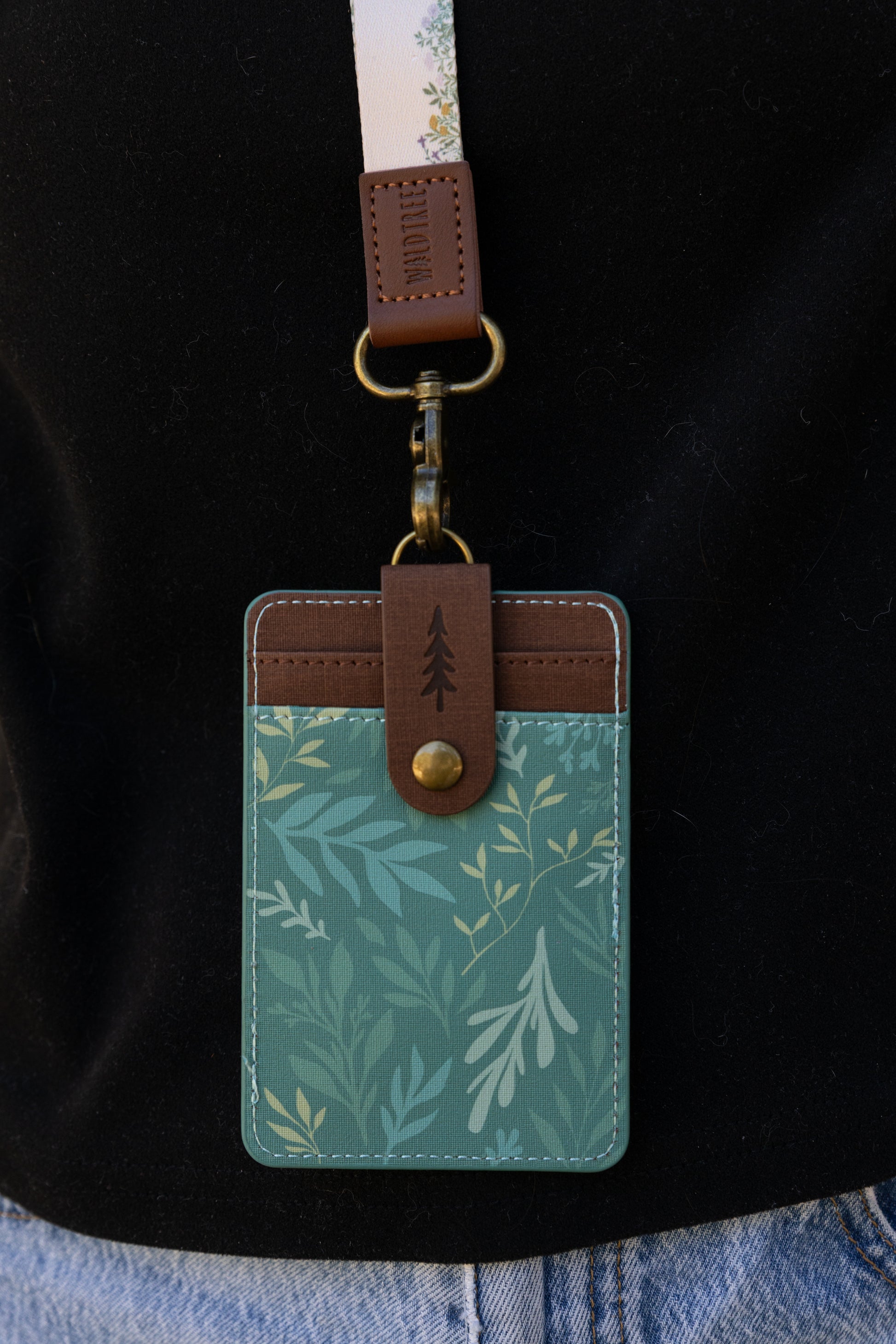 A close-up shot of the back of the green floral ID holder attached to a lanyard. It highlights the brown faux-leather snap strap with a debossed pine tree icon that secures the extra card slots.