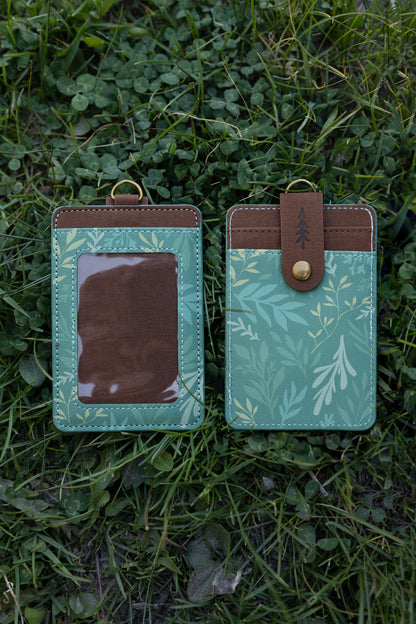 Front and back views of the green botanical ID holder laying flat on green grass. The photo displays the clear front viewing window on one side and the button-strap card slots on the other.