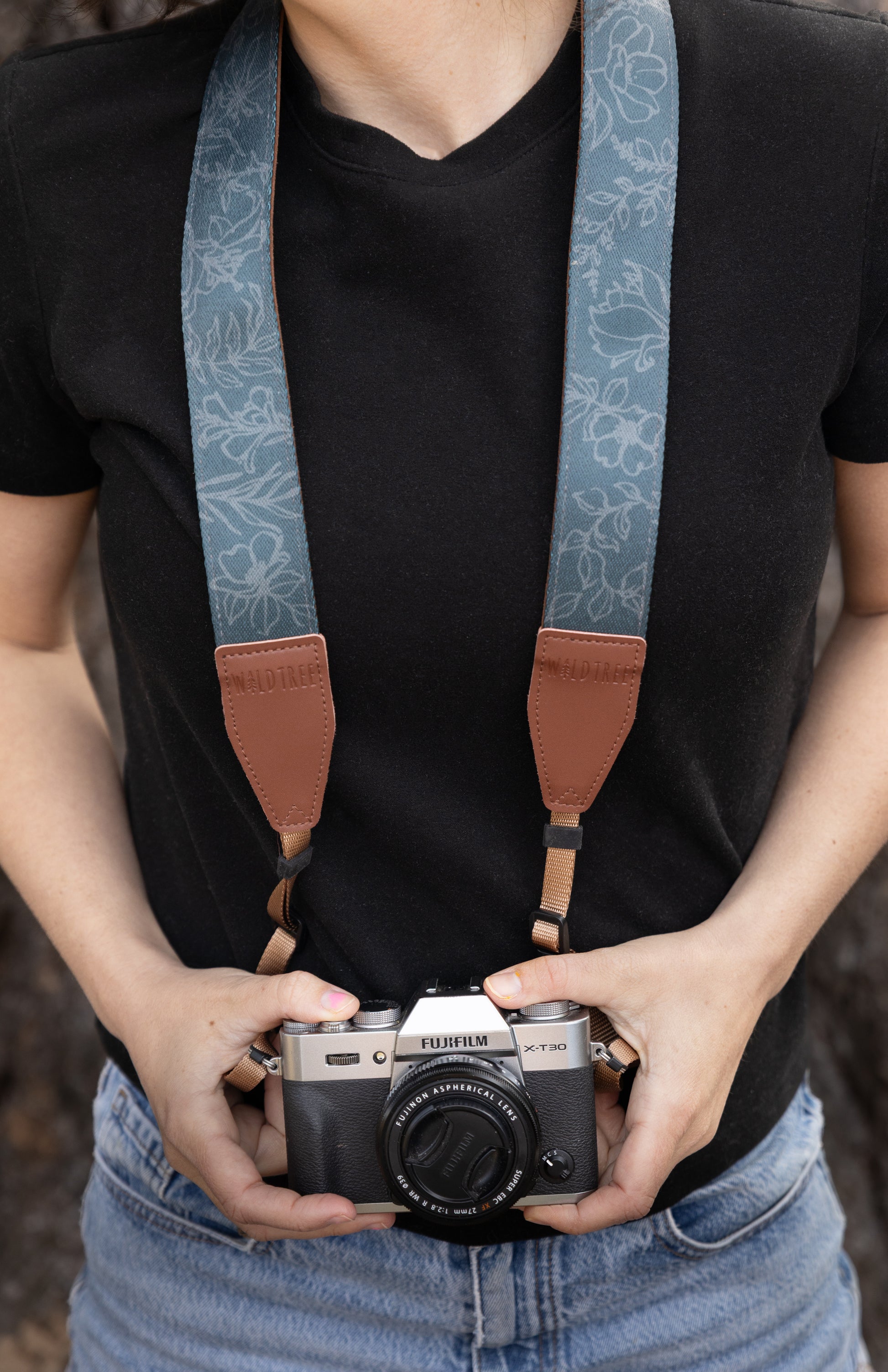 A front-facing lifestyle shot of a photographer holding a silver Fujifilm camera, featuring the Shadow Garden camera strap. The slate-blue strap displays a hand-drawn wildflower pattern and is secured with sturdy brown leather connectors branded with the WILDTREE logo.
