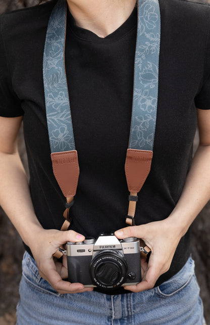 A front-facing lifestyle shot of a photographer holding a silver Fujifilm camera, featuring the Shadow Garden camera strap. The slate-blue strap displays a hand-drawn wildflower pattern and is secured with sturdy brown leather connectors branded with the WILDTREE logo.