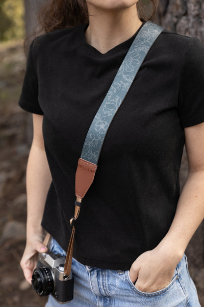 A lifestyle photograph showing the slate-blue floral camera strap worn cross-body by a photographer in an outdoor setting. The botanical design is paired with durable brown leather accents and a tan adjustable nylon cord attached to a silver mirrorless camera.