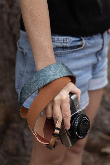 A close-up detail shot of the Shadow Garden camera strap wrapped around a photographer's wrist. The image highlights the contrast between the slate-blue floral patterned fabric and the soft, tan-colored underside, emphasizing the strap's premium materials and flexible design.